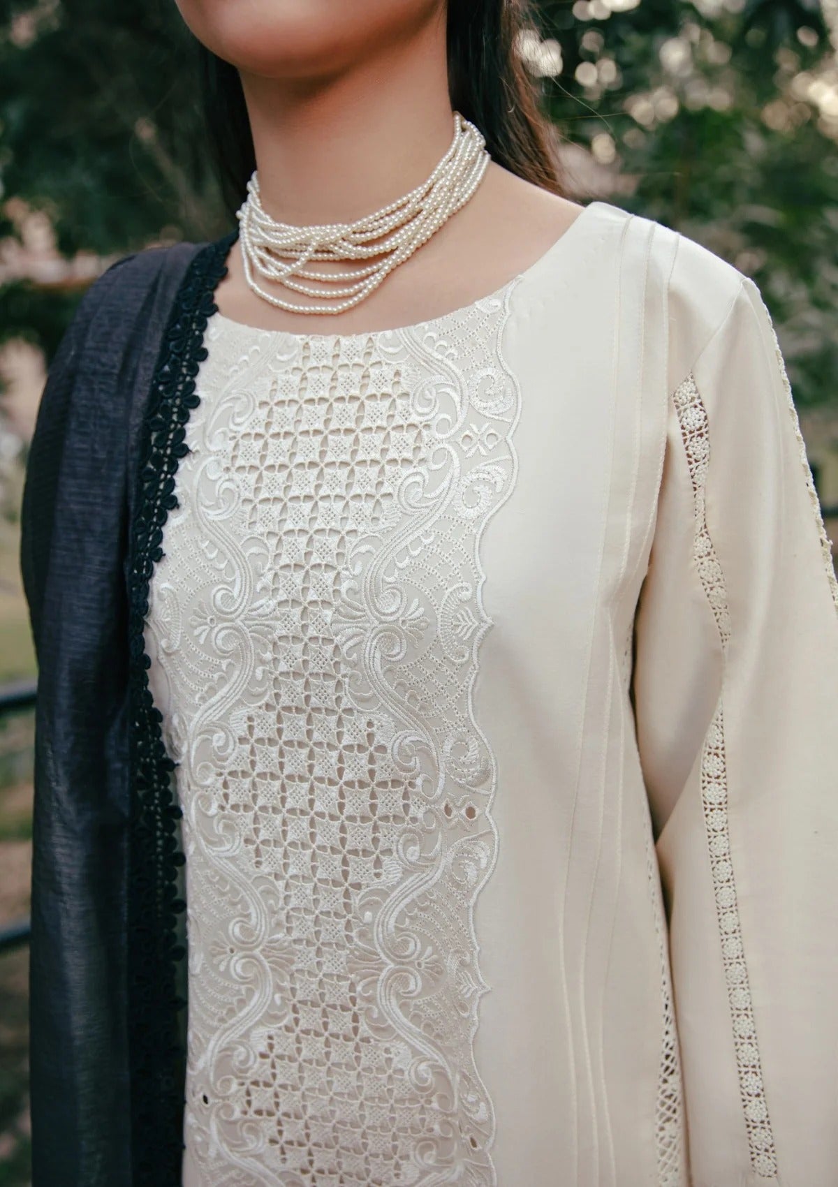 A close-up of a person wearing a cream-colored embroidered tunic with a matching black dupatta and a multi-strand pearl necklace, set against a blurred outdoor background.