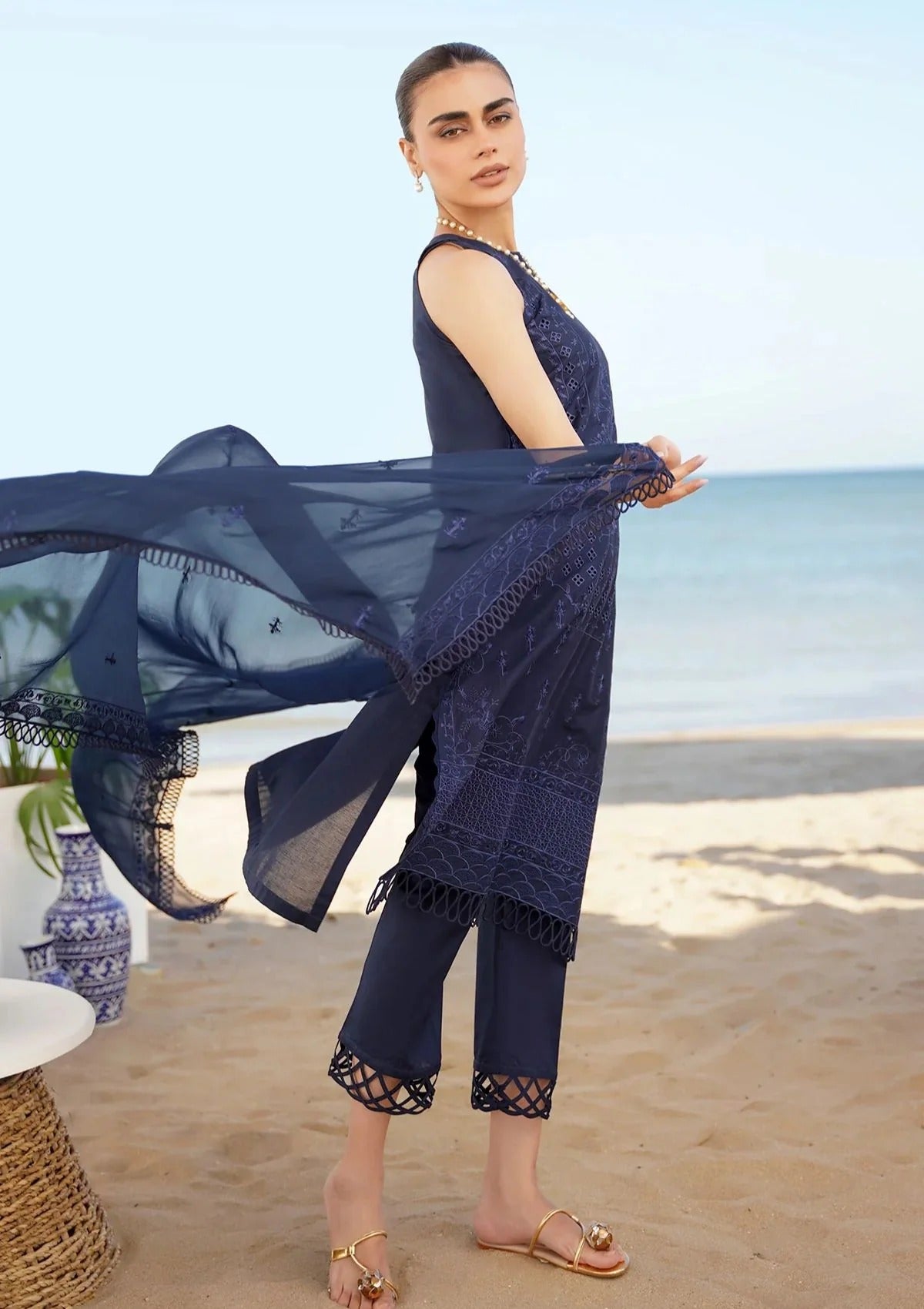A woman modeling a navy blue three-piece ethnic outfit with matching sheer dupatta on a sandy beach. The outfit features intricate lace embroidery, fringed details, and lace-up ankle cuffs.