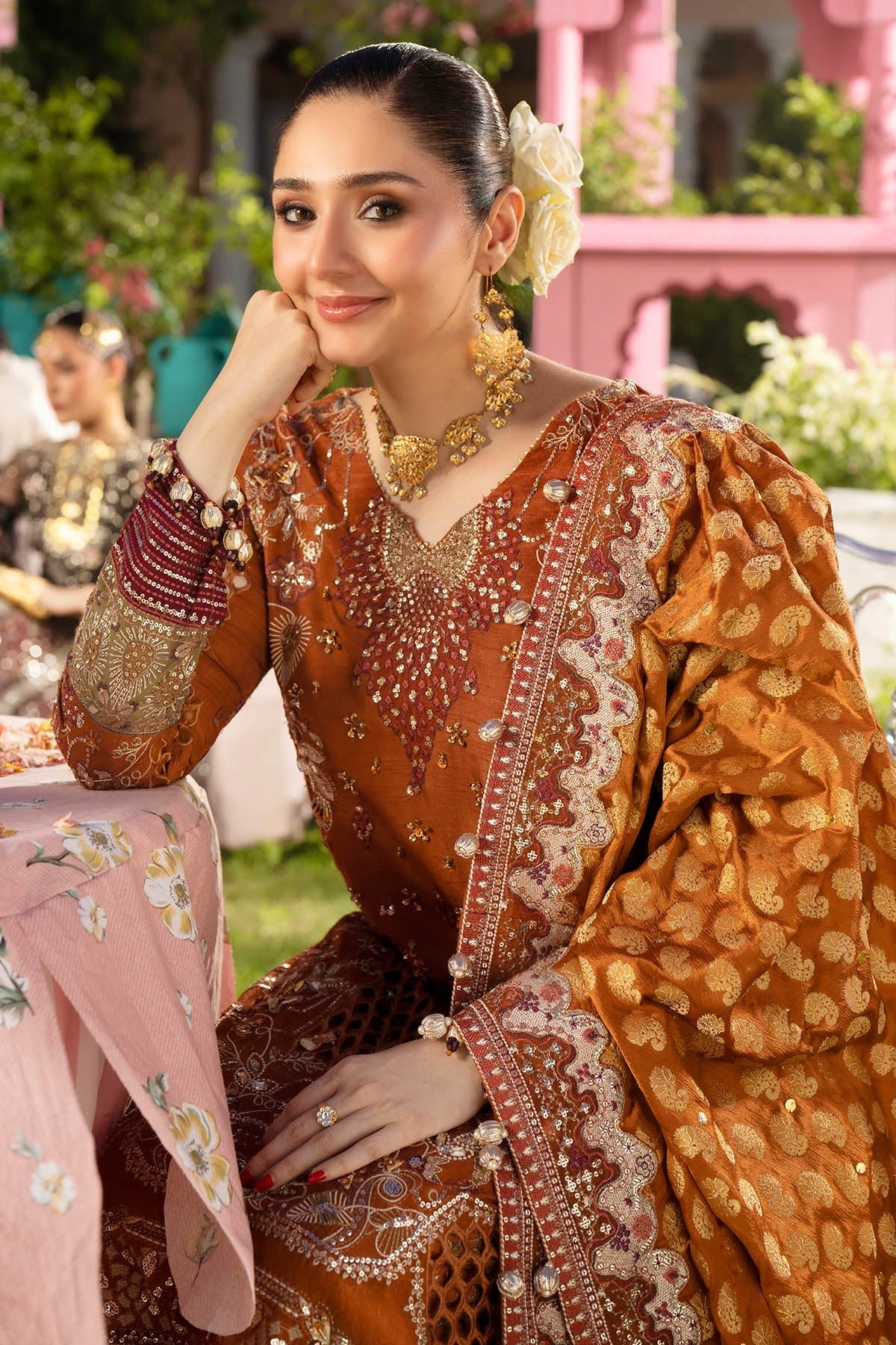 A woman wearing an ornate brown and gold embroidered traditional outfit with a pink floral dupatta, seated outdoors in front of a pink building.