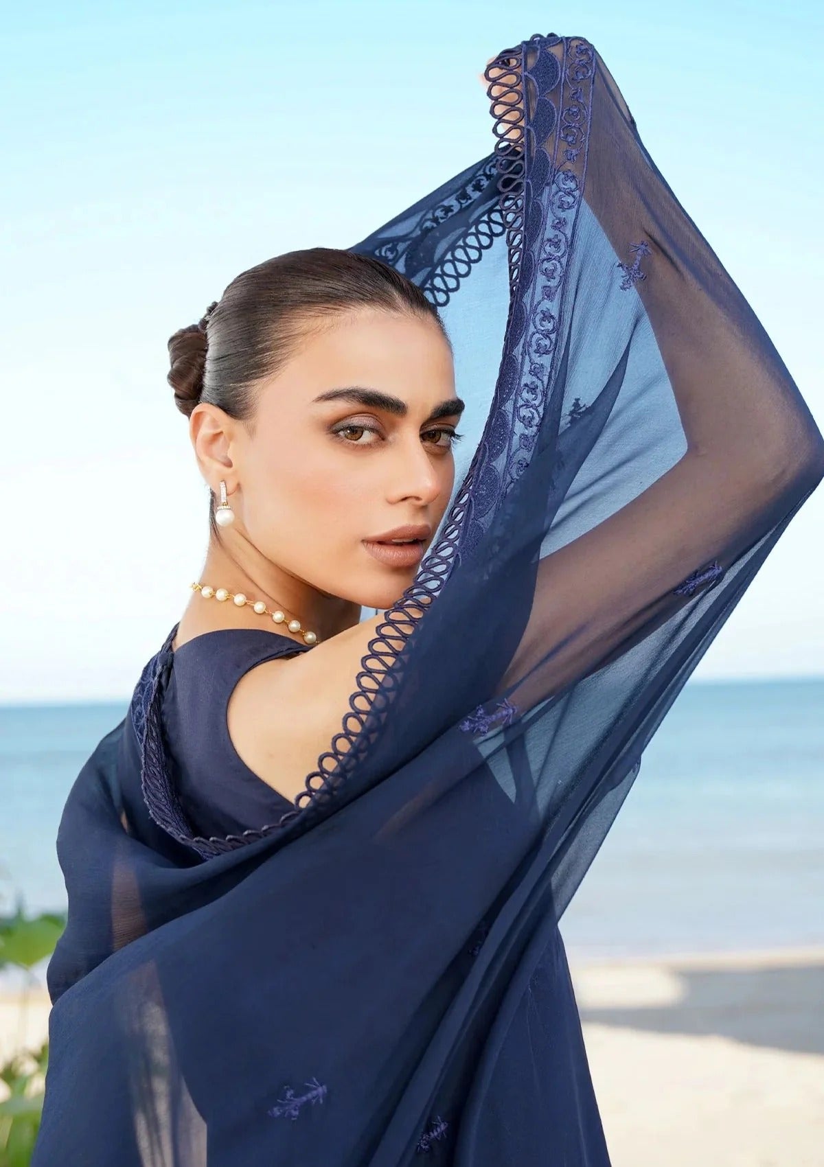 A woman wearing a navy blue sheer scarf with a decorative lace border, posing outdoors with the ocean and sky in the background.
