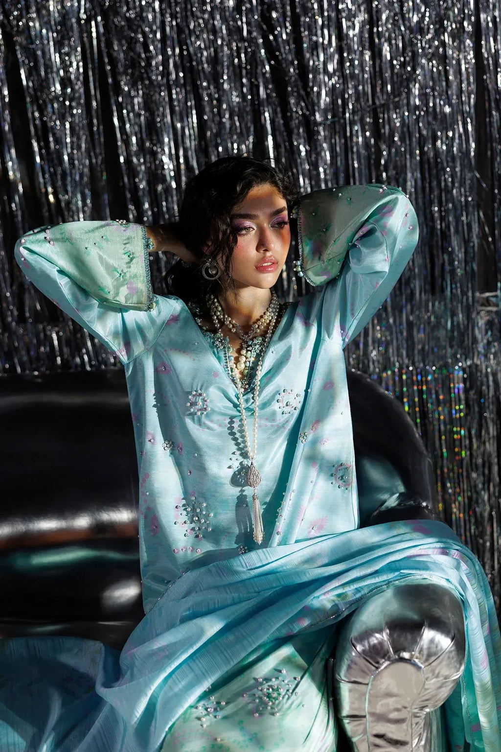 A woman wearing a flowing light blue embroidered tunic with metallic silver embellishments, seated against a shiny silver tinsel backdrop.