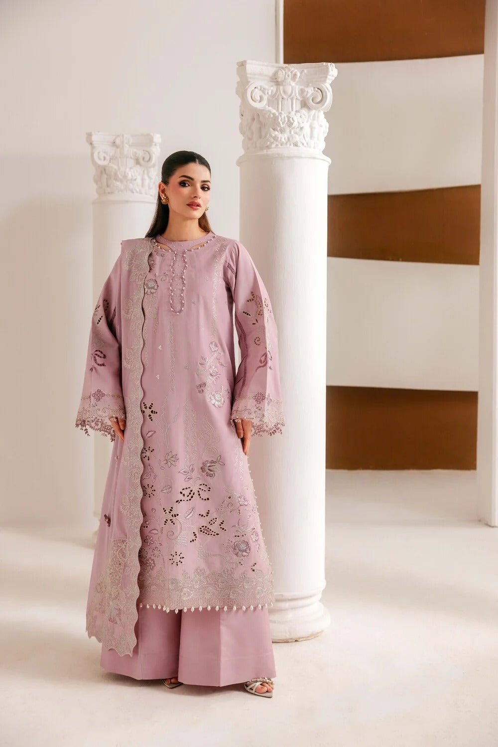 A woman modeling a light pink traditional South Asian outfit with floral embroidery and lace details, standing between white decorative columns in a minimalist indoor setting.