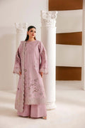 A woman modeling a light pink traditional South Asian outfit with floral embroidery and lace details, standing between white decorative columns in a minimalist indoor setting.