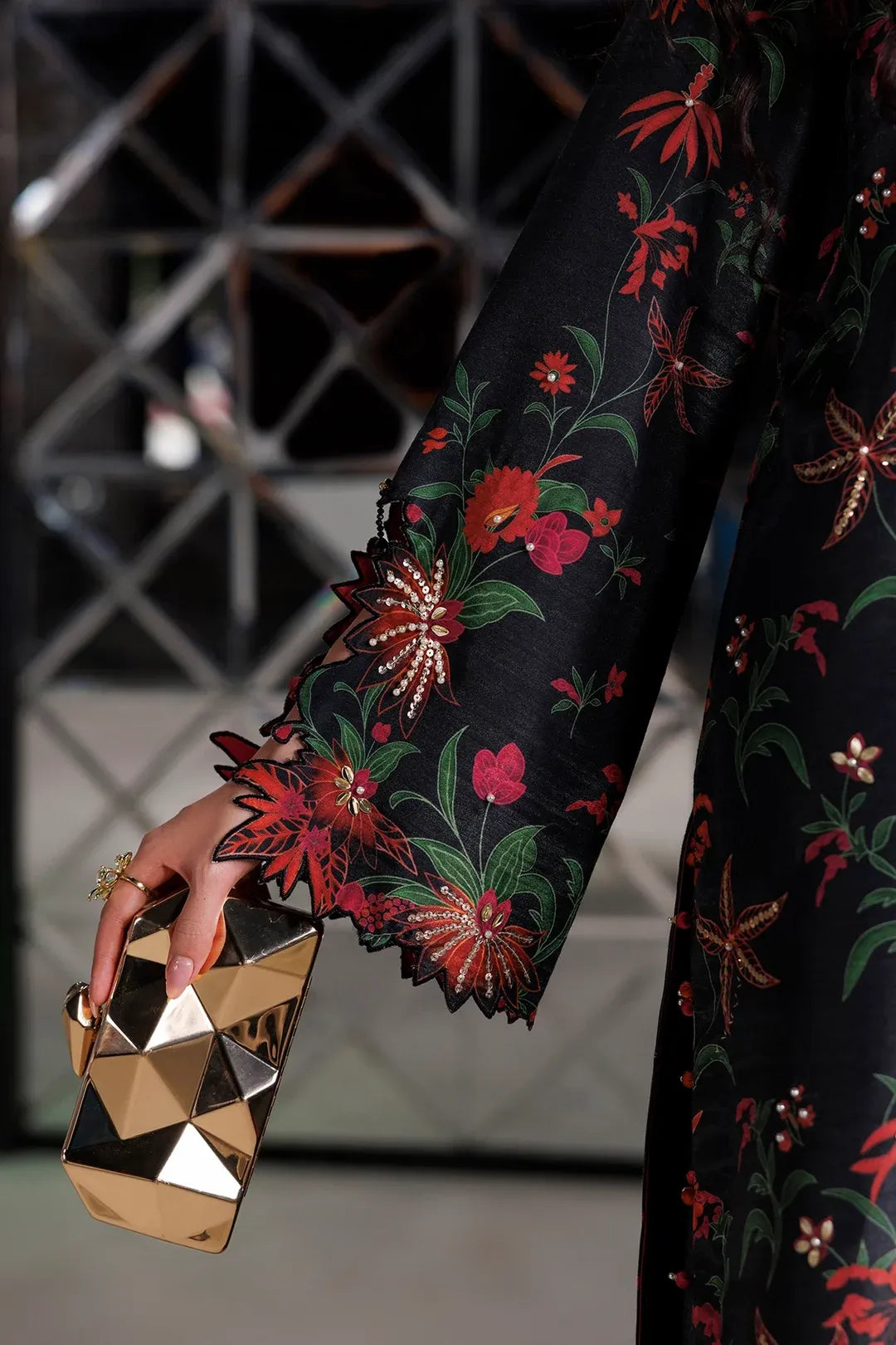A close-up of a person wearing a black long-sleeve dress with a vibrant red, green, and gold floral pattern, holding a faceted metallic gold clutch.