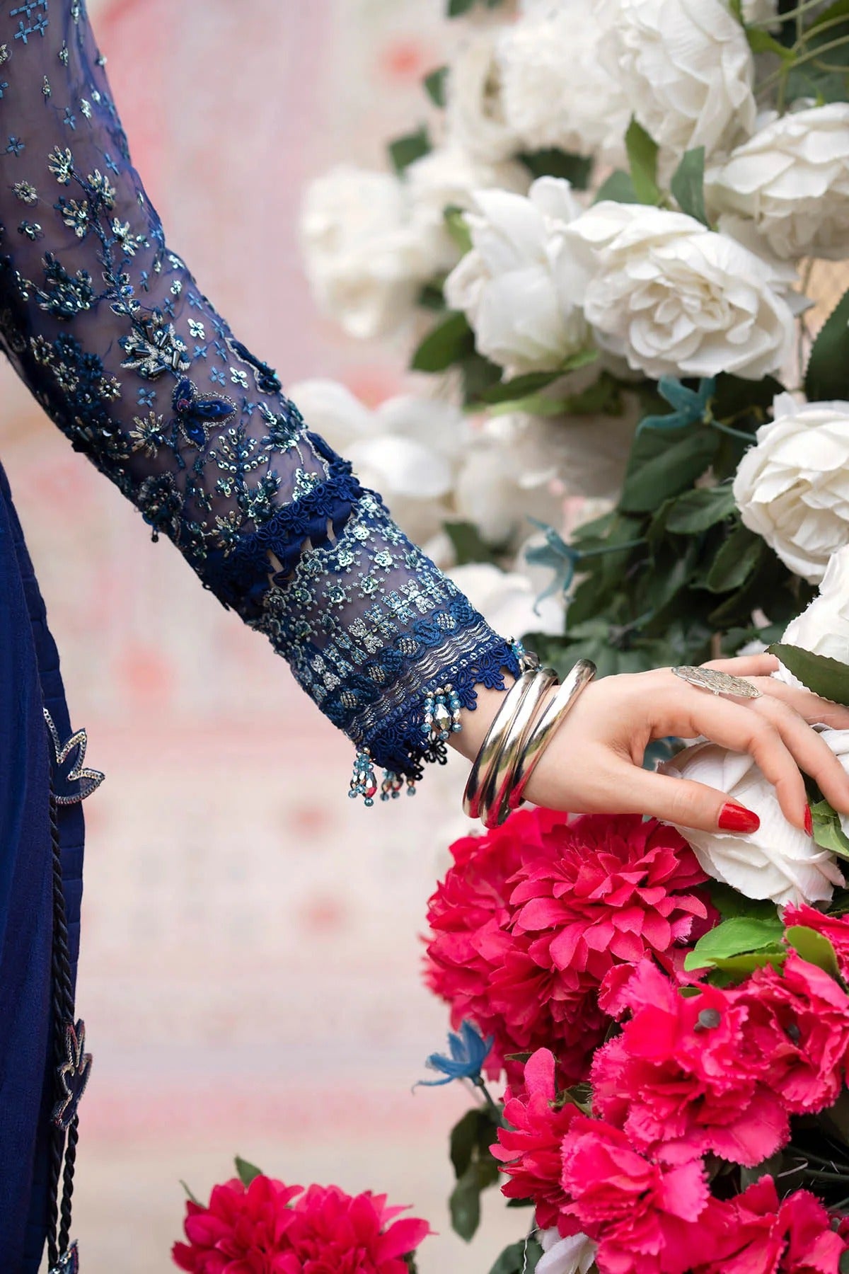 A close-up lifestyle shot of a person's hand adorned with stacked silver bangles and a ring, resting on a vibrant bouquet of white roses and bright pink peonies. The person is wearing a navy blue embroidered sleeve.