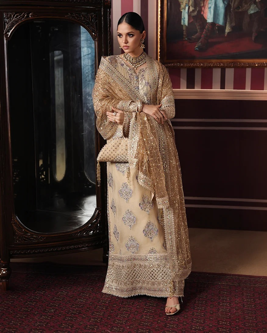 A woman models an elegant beige and gold embroidered traditional outfit with a matching sheer dupatta, standing in a richly decorated room with a large dark wooden mirror and red patterned carpet.