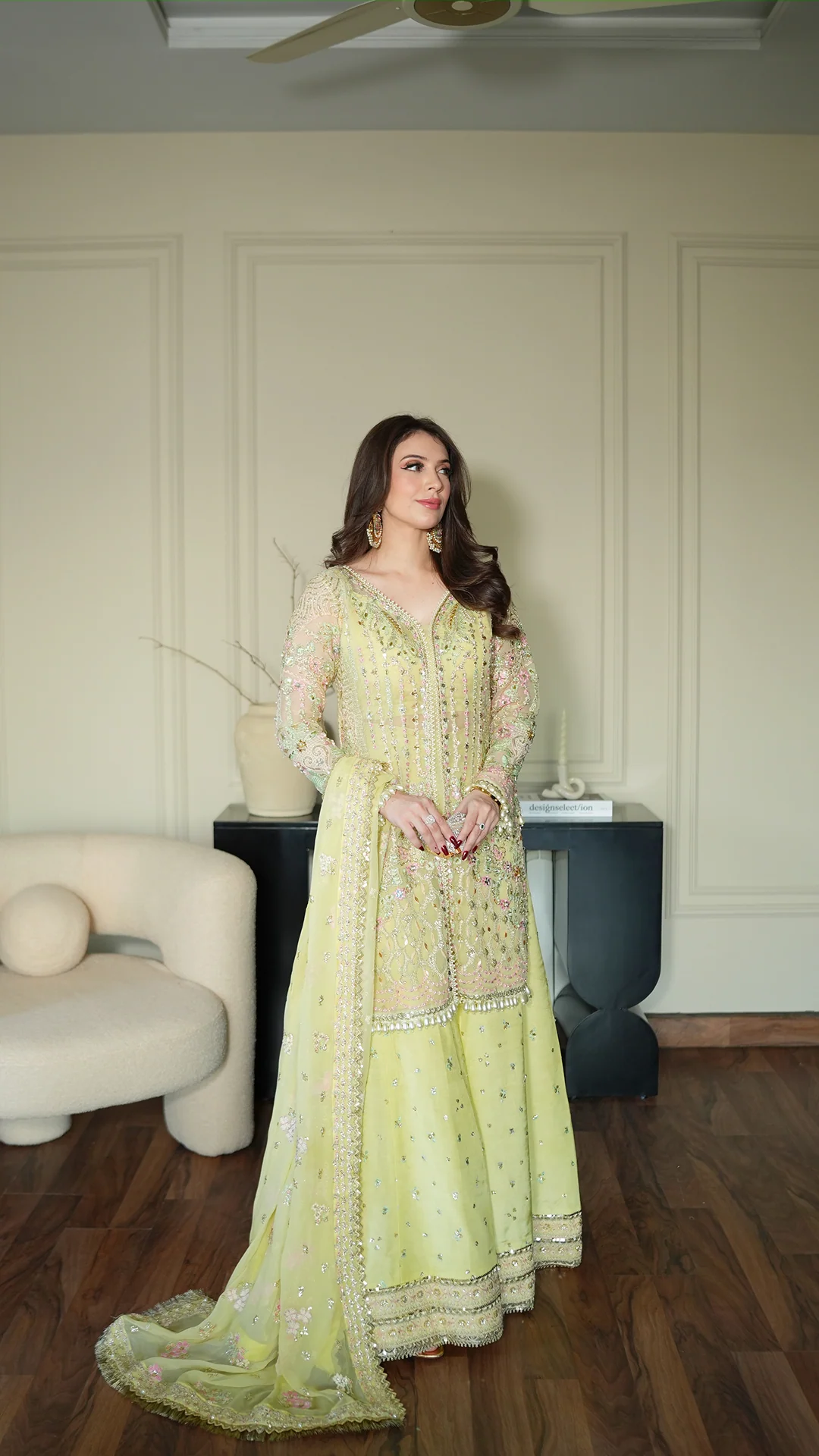 Woman in a light green traditional outfit standing in a room with a white wall and a chair.