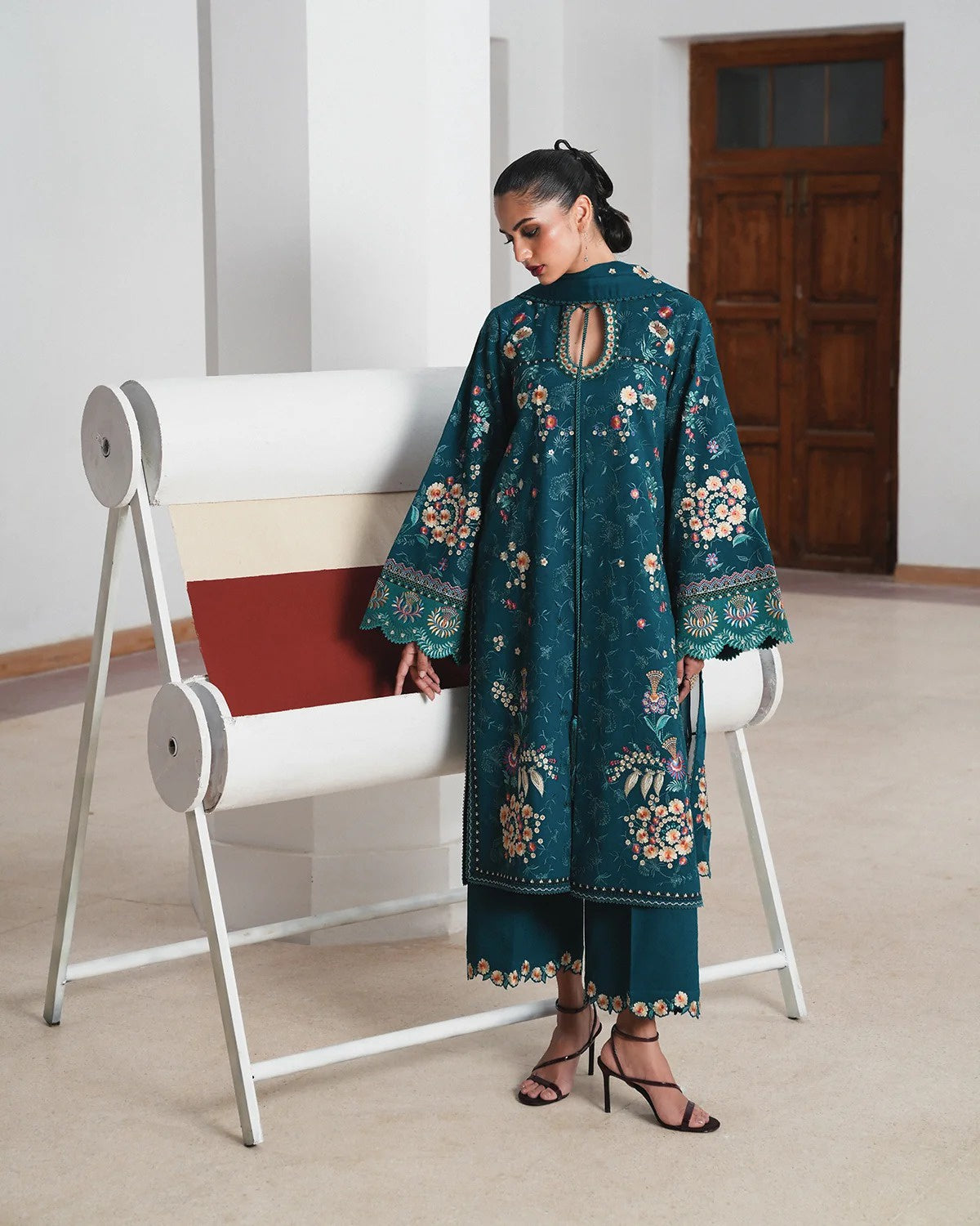A woman models a teal green embroidered kurta and matching trousers in a minimalist indoor setting with a white bench.