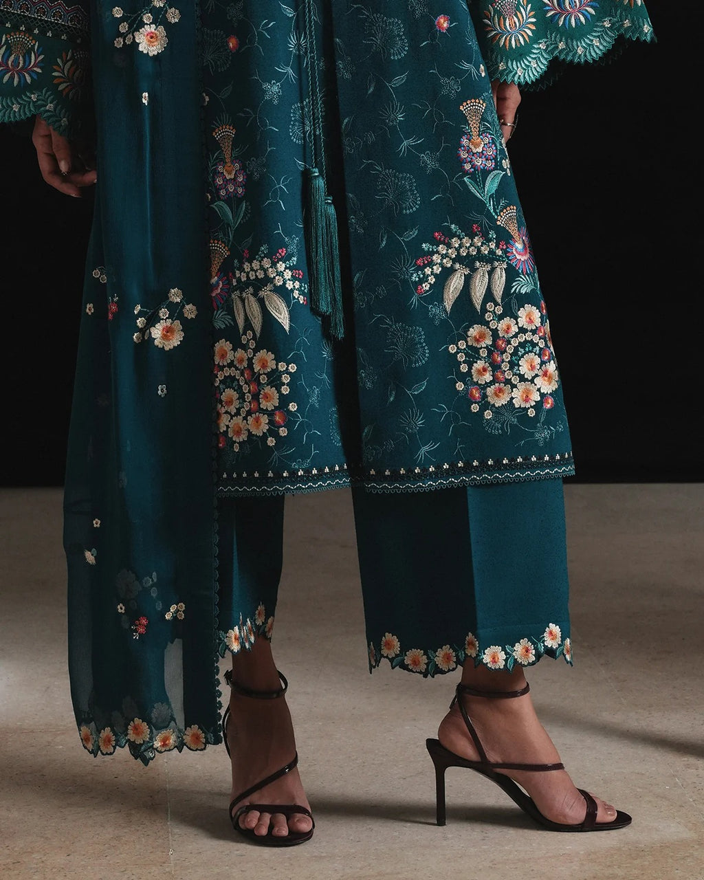 A close-up of a model wearing a teal green embroidered traditional outfit with floral patterns, paired with black strappy high-heeled sandals against a dark studio background.