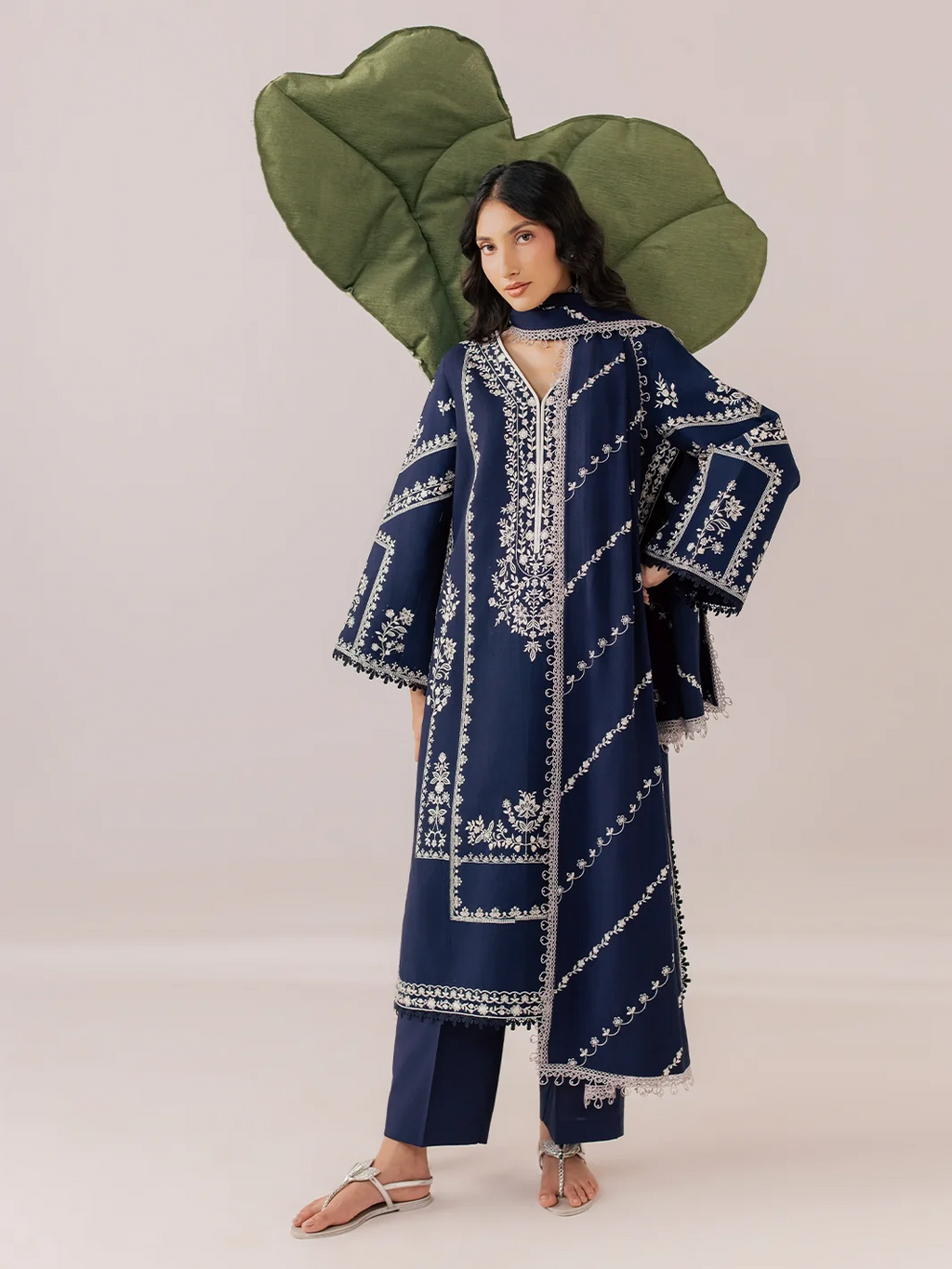 A model wearing a navy blue embroidered kaftan tunic with white patterns, matching navy pants, and a large olive green leaf-shaped headpiece against a solid light beige background.