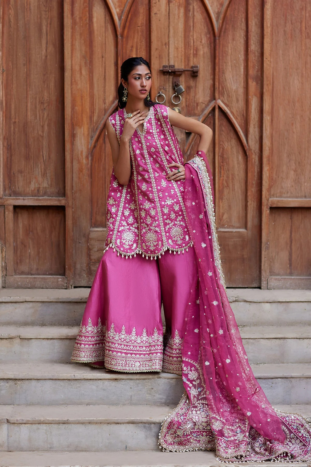 A woman models a vibrant pink traditional South Asian outfit with intricate gold embroidery, wide-leg pants, and a matching dupatta, posed in front of a rustic wooden door and stone steps.