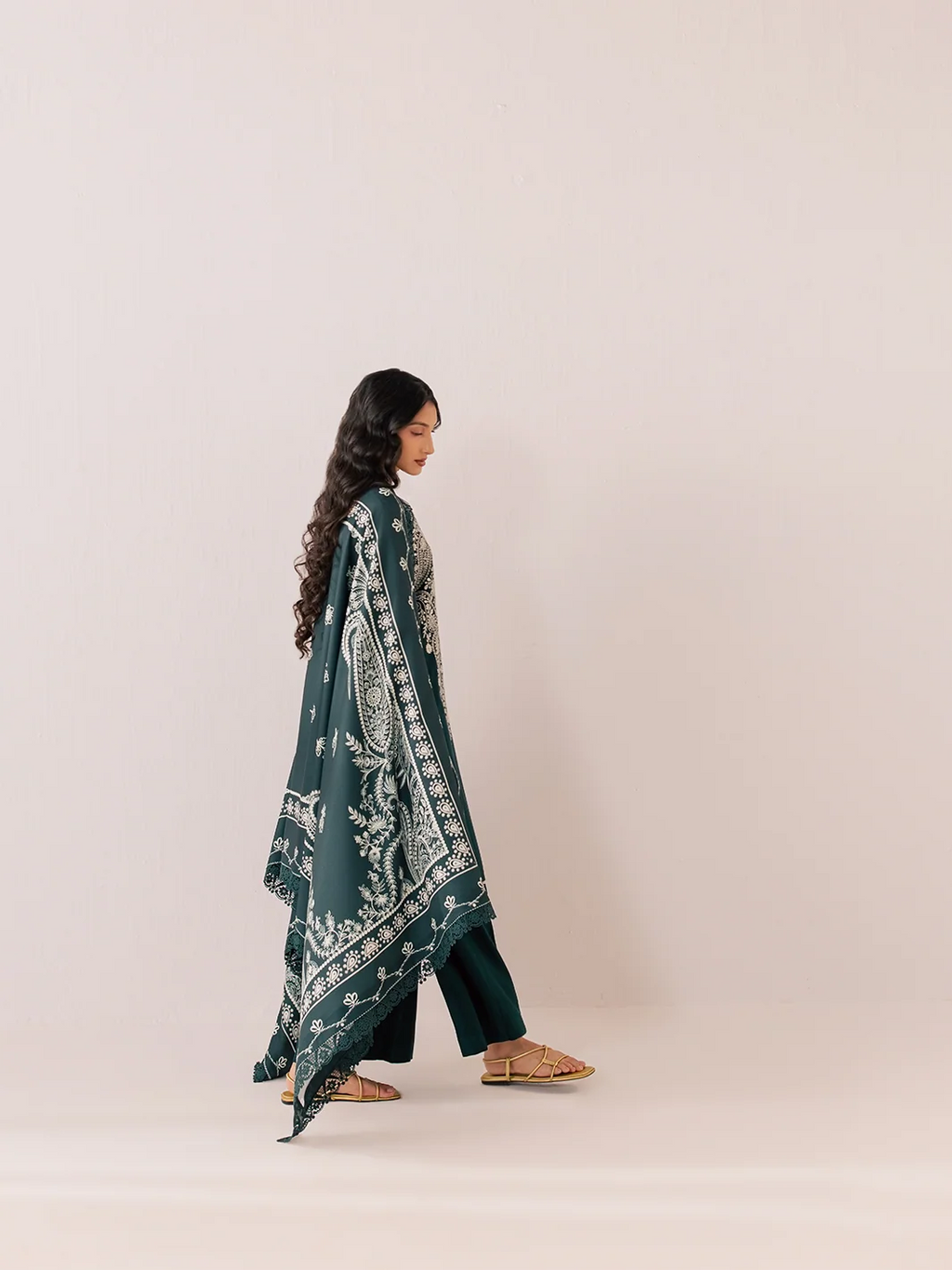 A woman is shown from the side wearing a dark teal green traditional outfit with a matching printed dupatta and dark green pants, standing against a solid light beige background.