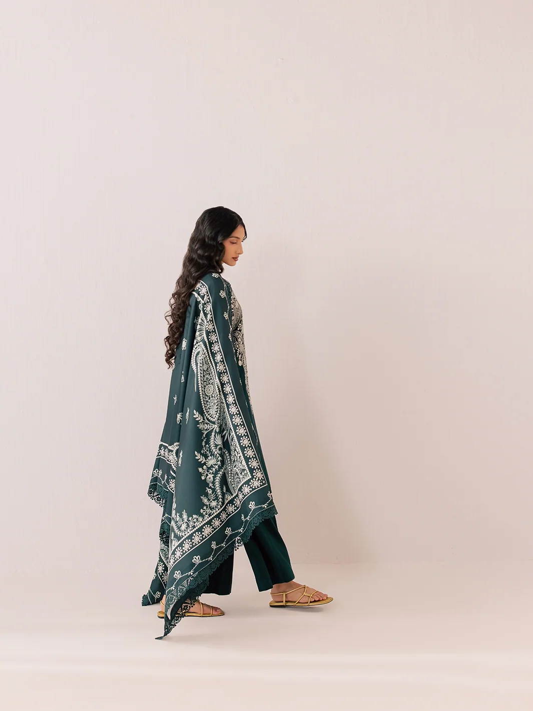 A woman is shown from the side wearing a dark teal green traditional outfit with a matching printed dupatta and dark green pants, standing against a solid light beige background.