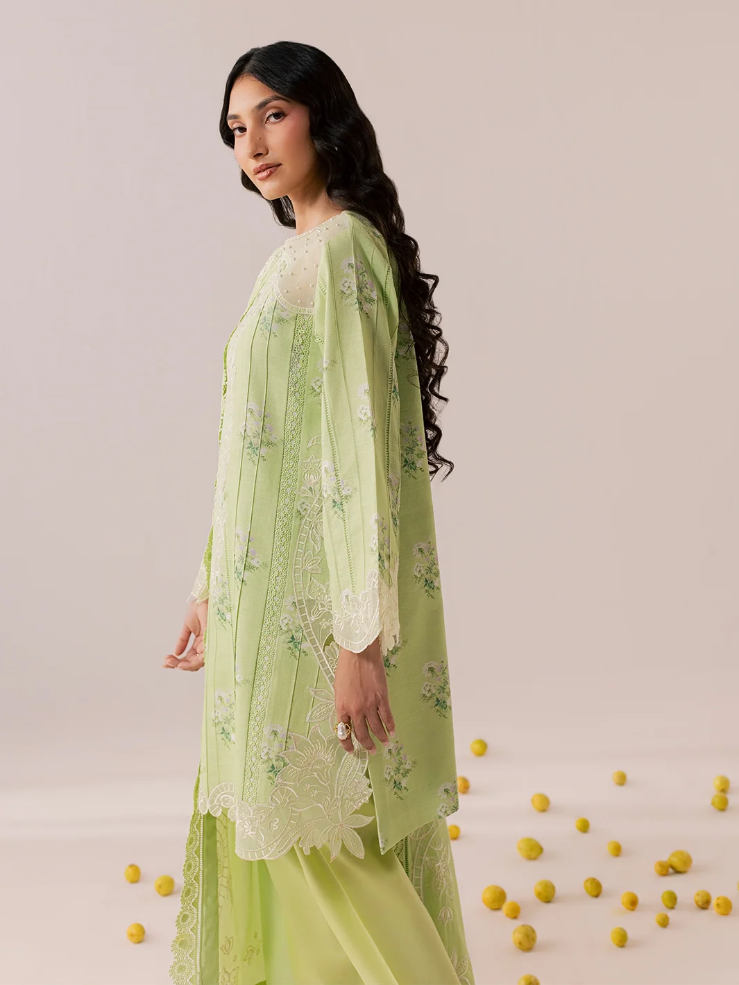 A woman models a light green traditional outfit with floral embroidery and lace details, photographed against a soft beige studio backdrop with scattered yellow flower petals on the floor.