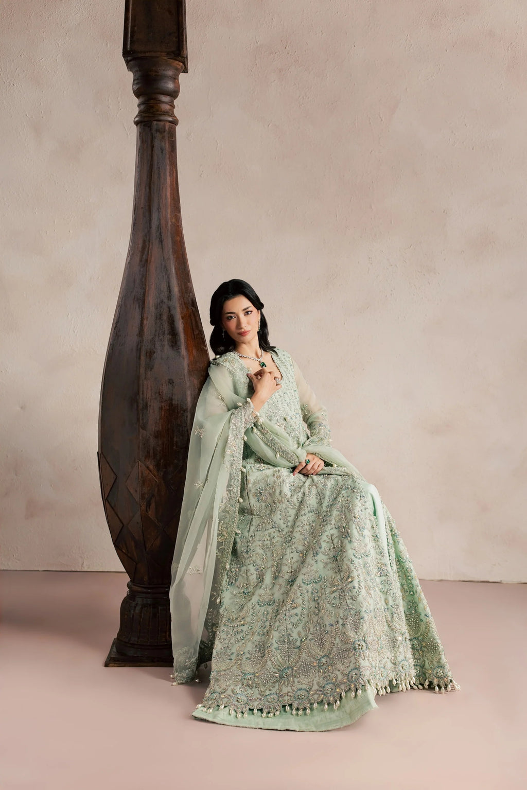A woman wearing a light mint green embroidered traditional outfit with a matching dupatta, seated beside a large dark wooden pillar against a soft beige studio backdrop.