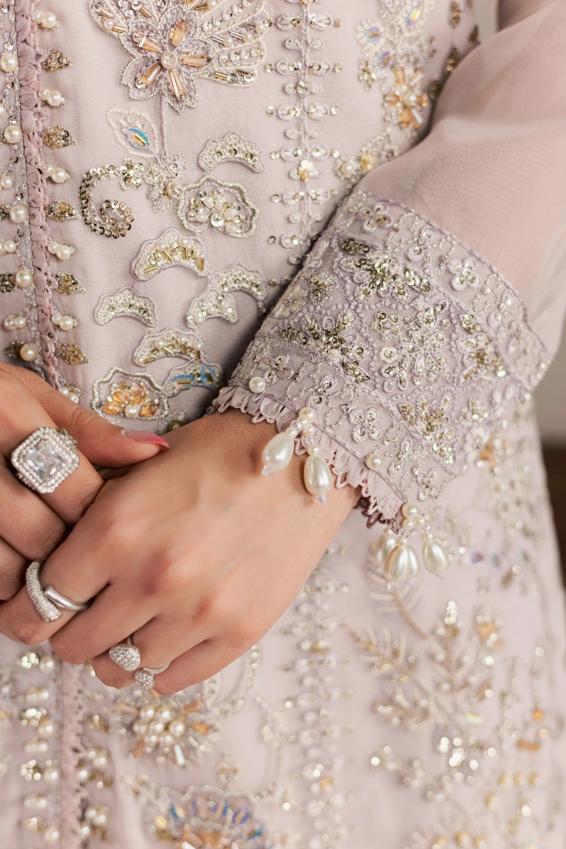 Close-up of a person wearing an ornate light pink embroidered traditional outfit with intricate silver, gold, and pearl beadwork, layered rings, and a pearl bracelet.