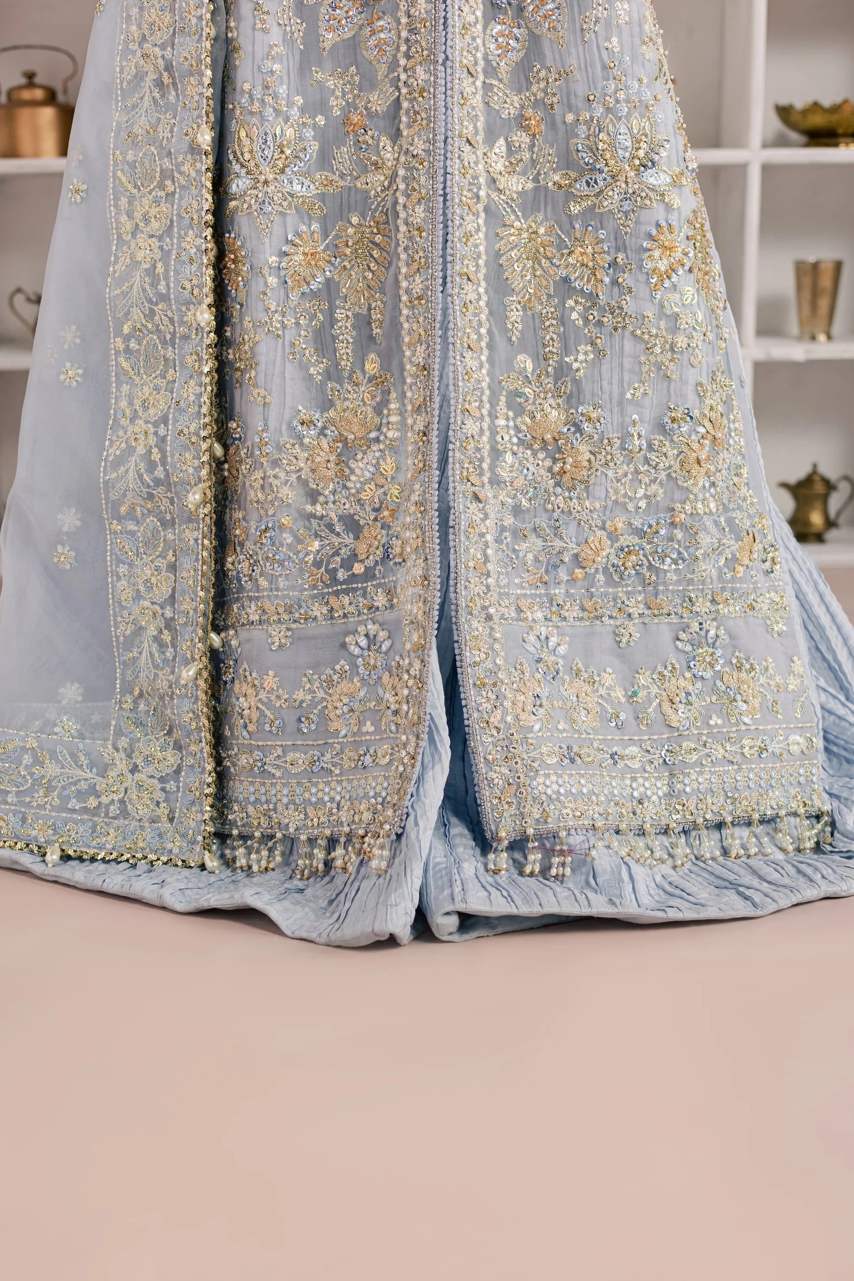 A close-up of a light blue traditional embroidered garment with ornate gold floral embroidery and beadwork, draped against a soft beige background with blurred shelf decor.