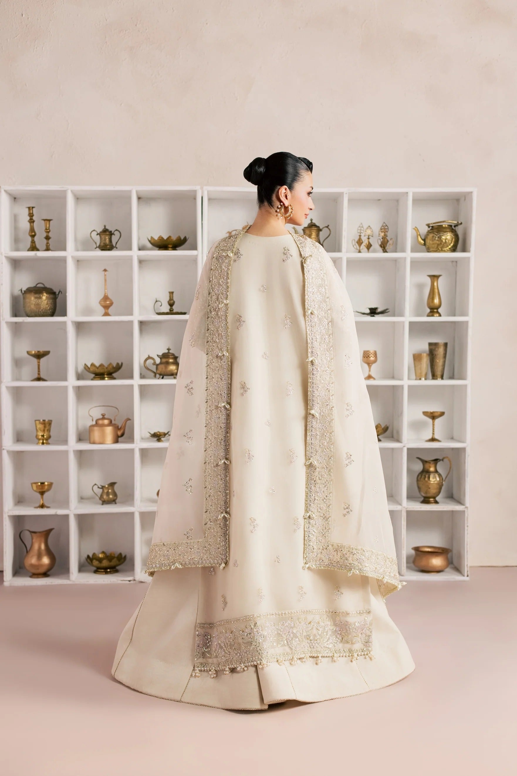 A woman stands with her back to the camera wearing a cream-colored traditional outfit with a sheer embroidered cape. She is positioned in front of a large white shelving unit filled with decorative brass and metallic objects against a soft beige wall.