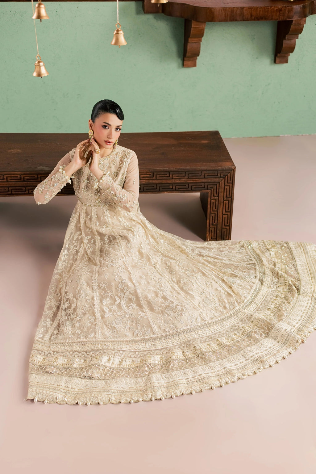 A woman models a long-sleeved cream-colored embroidered anarkali gown with a matching dupatta, posed in front of a dark wooden bench against a sage green wall.