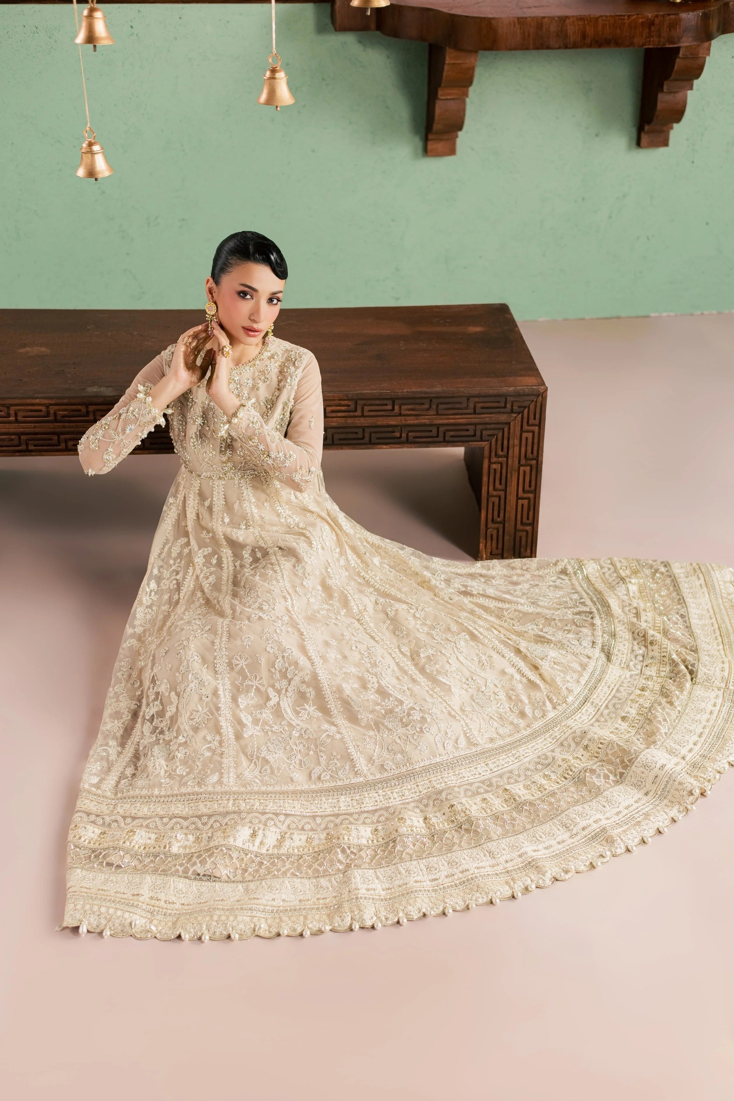 A woman models a long-sleeved cream-colored embroidered anarkali gown with a matching dupatta, posed in front of a dark wooden bench against a sage green wall.