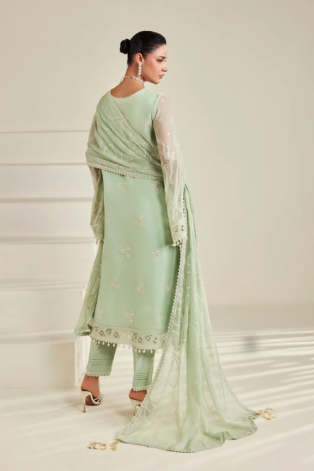 A woman modeling a light sage green traditional outfit with matching pants and a sheer dupatta, photographed in a clean studio setting with a cream background.