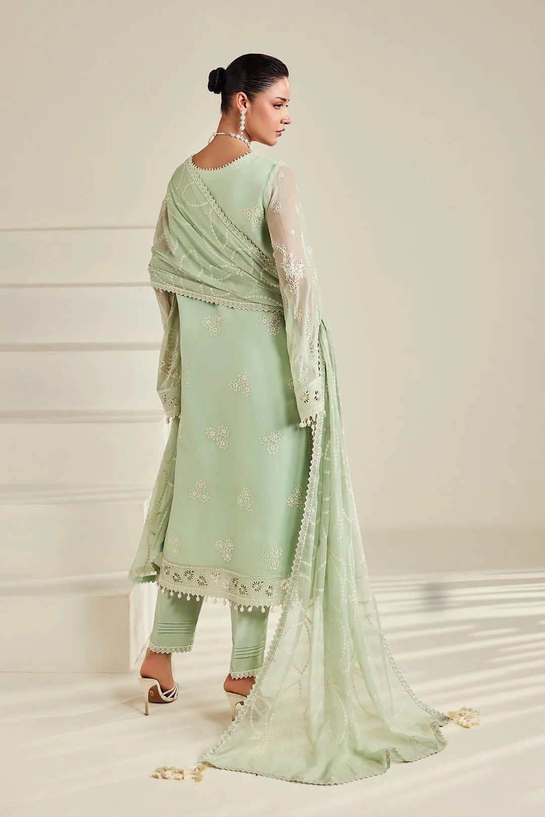 A woman modeling a light sage green traditional outfit with matching pants and a sheer dupatta, photographed in a clean studio setting with a cream background.