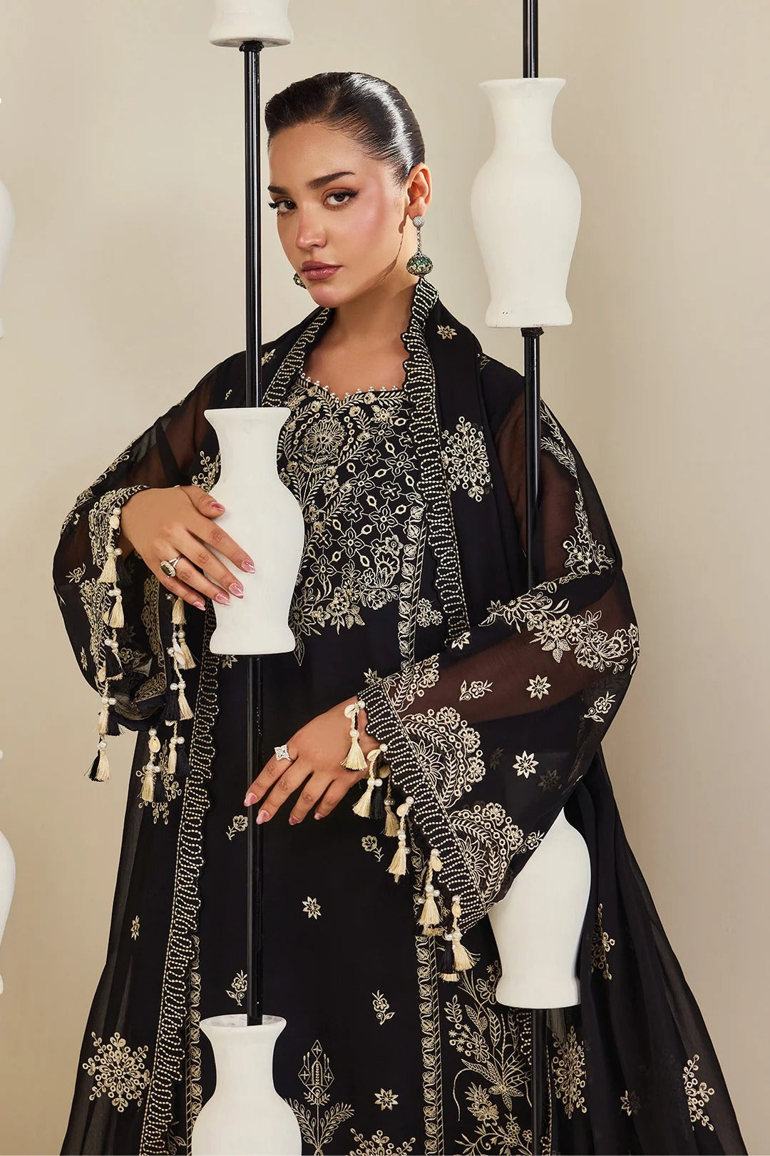 A woman models a black embroidered traditional outfit with sheer sleeves and gold-toned tassel details, posed among white decorative vases against a beige studio background.