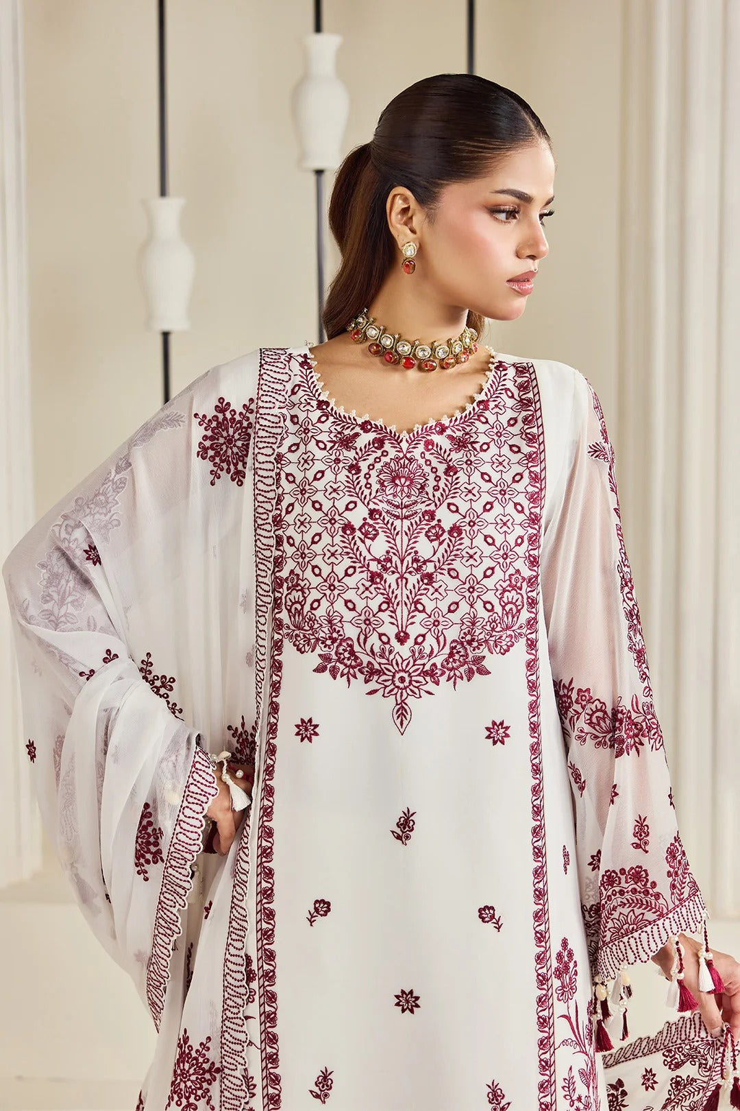 A woman modeling a traditional white ethnic kurta set with deep maroon embroidery and a matching sheer embroidered dupatta, set against a soft beige background with hanging decorative vases.