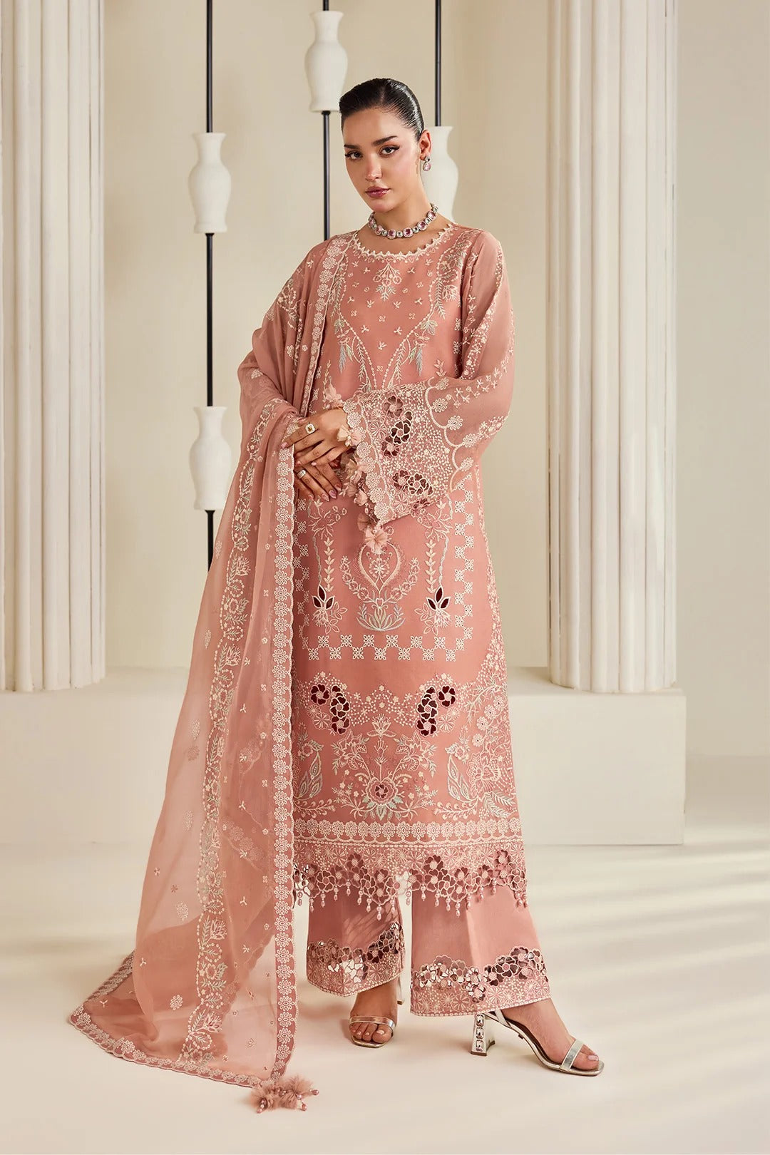 A woman models a peach-pink traditional embroidered outfit with a matching dupatta, featuring intricate silver and black embroidery, set against a classical cream-colored architectural background.