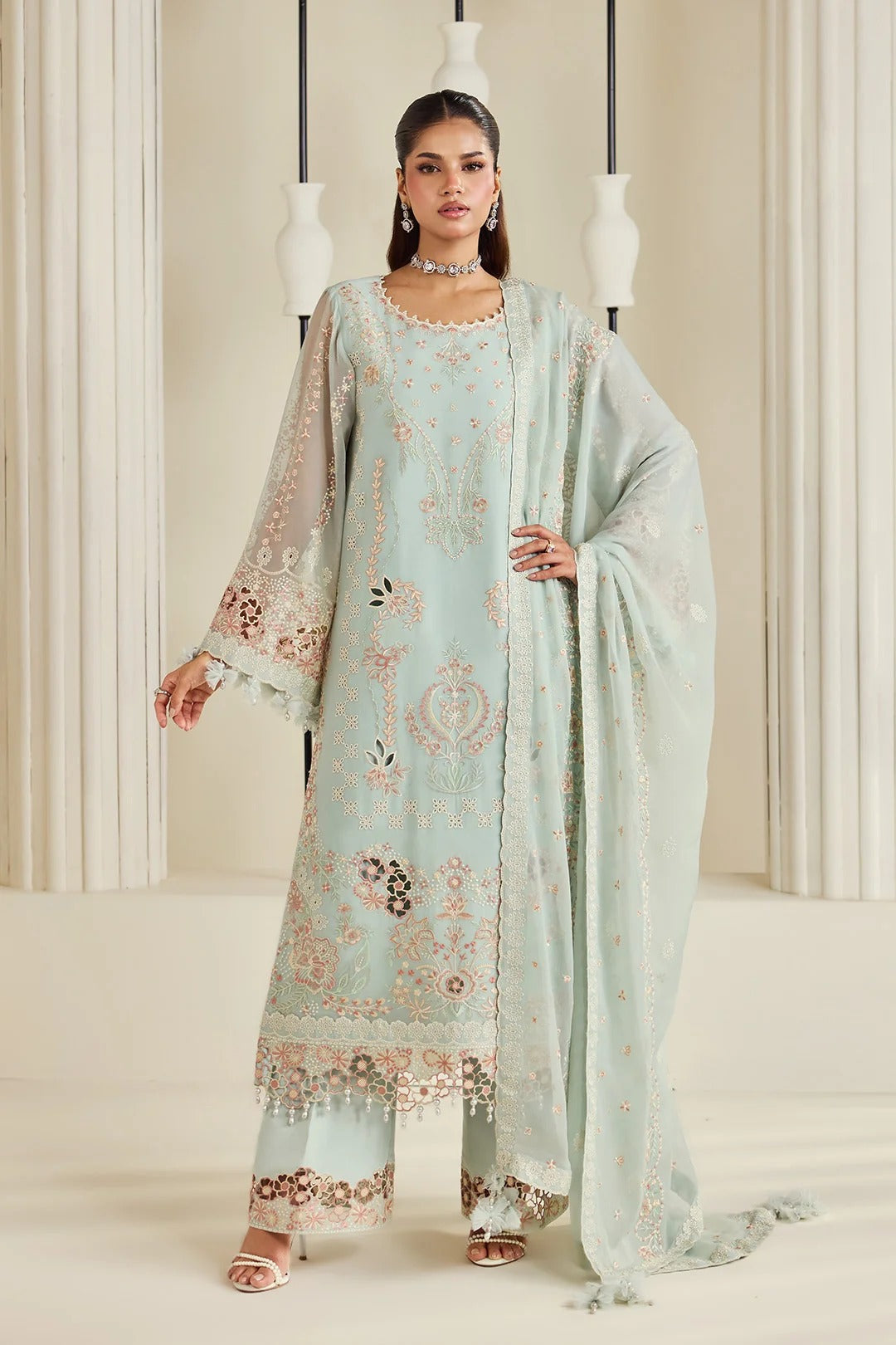 A woman models a traditional light blue-green salwar kameez suit with intricate floral embroidery in beige and brown, paired with a matching sheer dupatta, set against a warm beige background with hanging white vases.