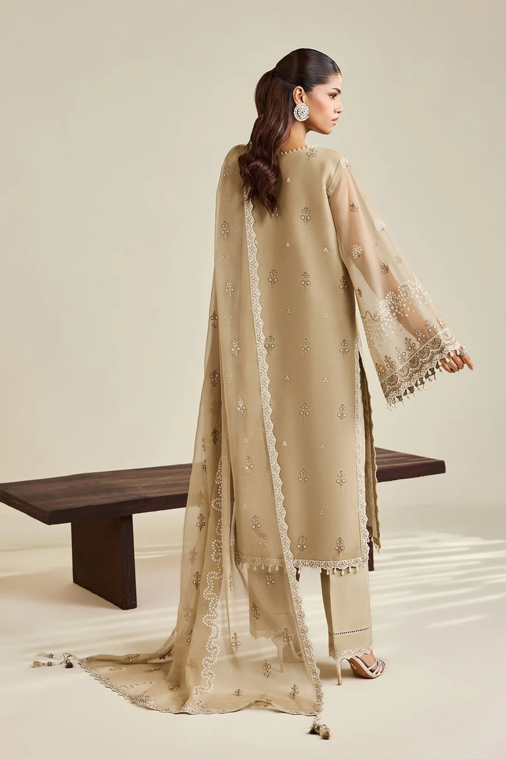 A woman modeling a beige traditional outfit with a matching sheer dupatta draped over her shoulders, photographed in a studio setting with a wooden bench in the background.