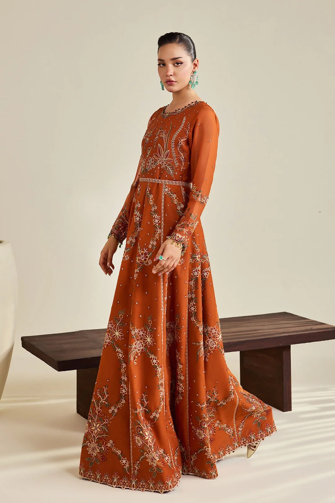 A woman models a long-sleeved rust-orange maxi dress with intricate silver and gold floral embroidery, standing in front of a minimalist beige background with a dark wooden bench.
