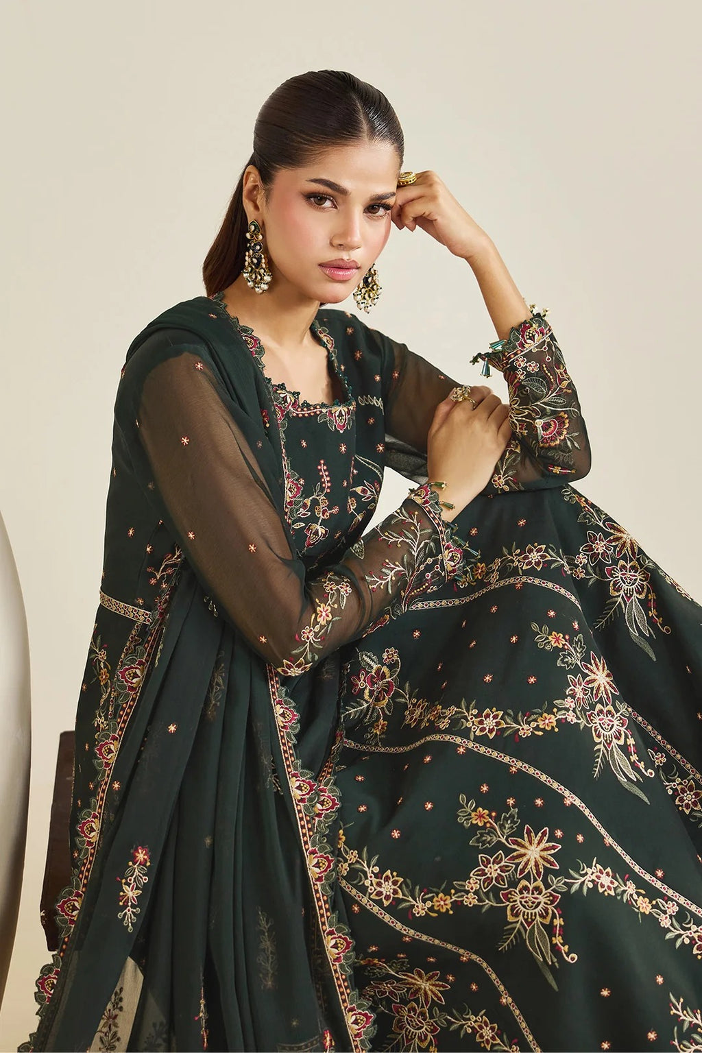 A woman models a dark green embroidered traditional outfit with sheer sleeves and a matching dupatta, decorated with intricate floral patterns in gold, pink, and red, set against a plain beige background.