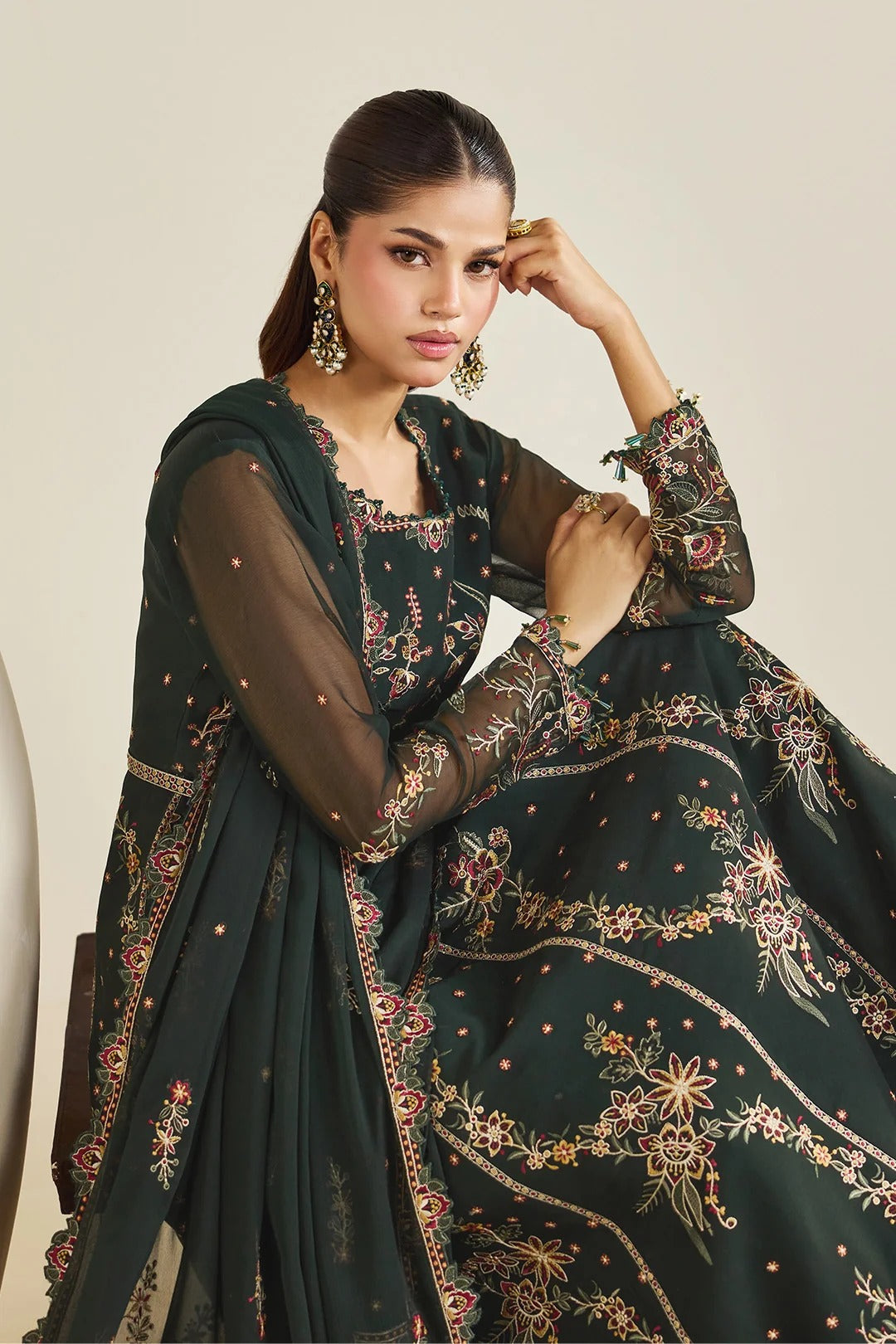 A woman models a dark green embroidered traditional outfit with sheer sleeves and a matching dupatta, decorated with intricate floral patterns in gold, pink, and red, set against a plain beige background.