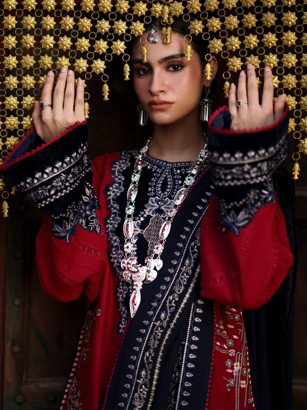 A woman wearing an ornate traditional outfit with a red and navy embroidered tunic, heavy silver jewelry, and a gold beaded curtain backdrop.