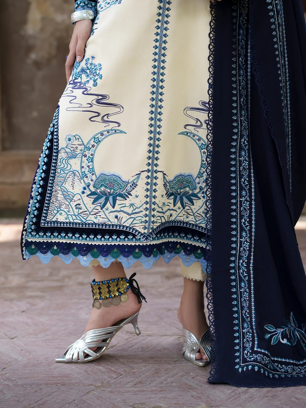 A close-up of a traditional South Asian outfit featuring a cream-colored kurta with intricate blue and green floral embroidery, paired with a navy blue dupatta and silver heeled sandals.
