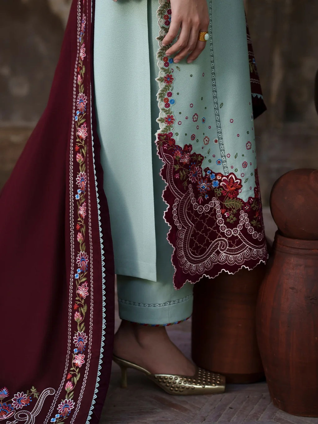 A close-up of a person wearing traditional South Asian attire, featuring a mint green kurta with intricate red and gold floral embroidery and a deep maroon dupatta with matching embroidery, styled with gold pointed-toe heels.