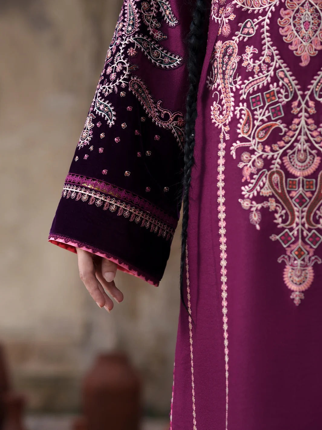 A close-up of a traditional embroidered garment in rich magenta-purple fabric, featuring ornate paisley and floral embroidery in pink, white, and gold, with a dark purple sleeve decorated with metallic embroidery and a hand resting near the cuff.