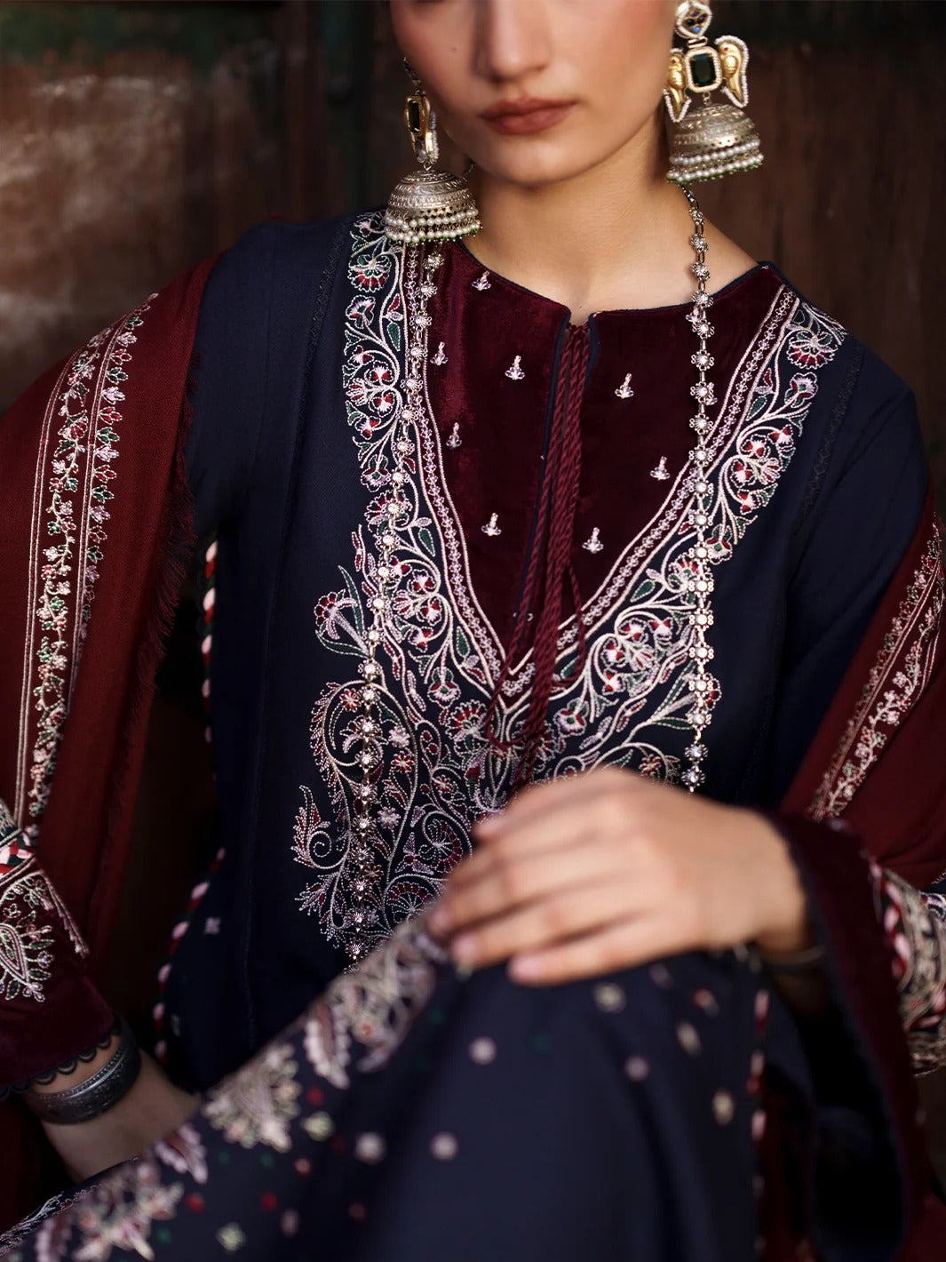 A close-up of a model wearing a traditional South Asian outfit featuring a navy blue kurta with intricate silver and gold embroidery, a deep maroon velvet inner layer, and a matching maroon dupatta.