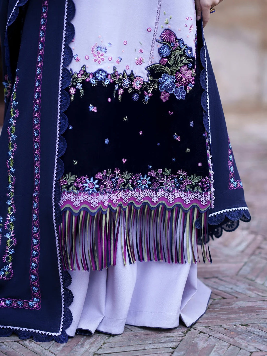 Close-up of a traditional embroidered garment with a navy blue border, multicolored floral embroidery, and long fringe, worn over a white dress on a person standing on a brick surface.