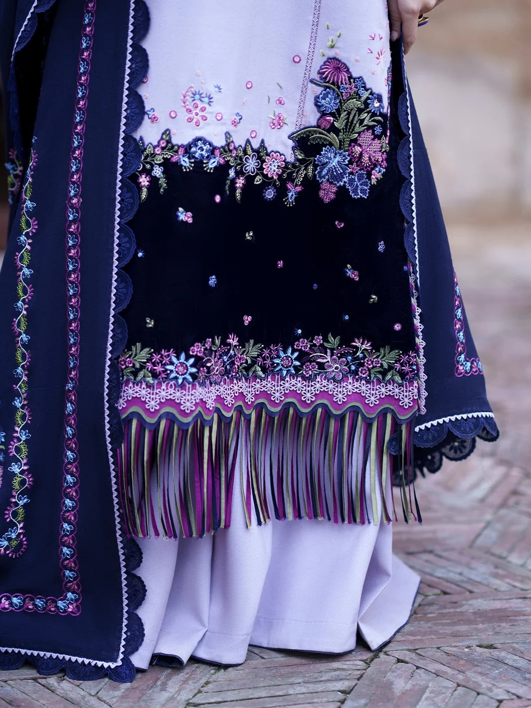 Close-up of a traditional embroidered garment with a navy blue border, multicolored floral embroidery, and long fringe, worn over a white dress on a person standing on a brick surface.