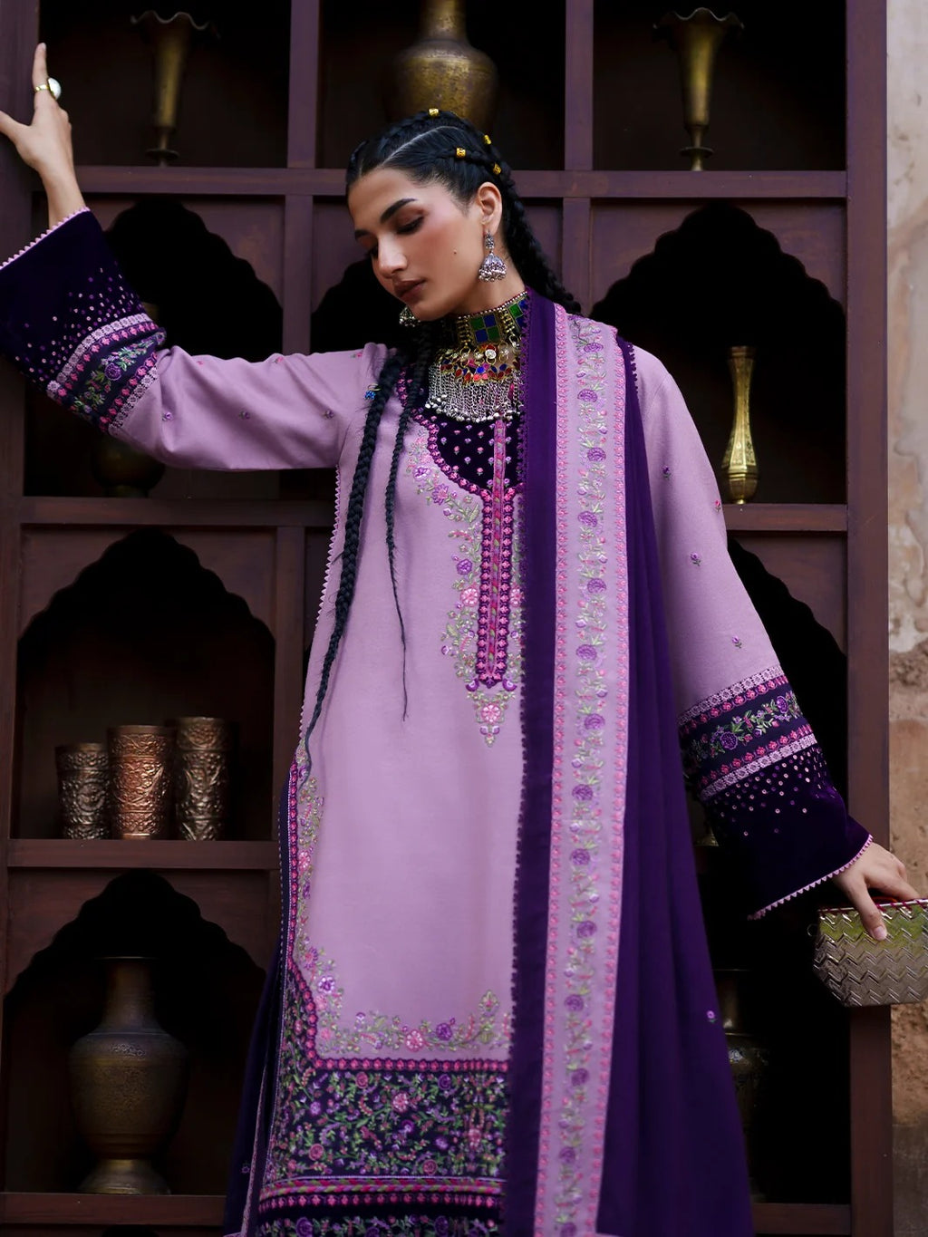 A woman models a traditional South Asian outfit featuring a lavender kurta with purple and gold embroidery, a matching purple dupatta, and a dark purple embroidered jacket, posed in front of a dark wooden shelf with decorative vases.