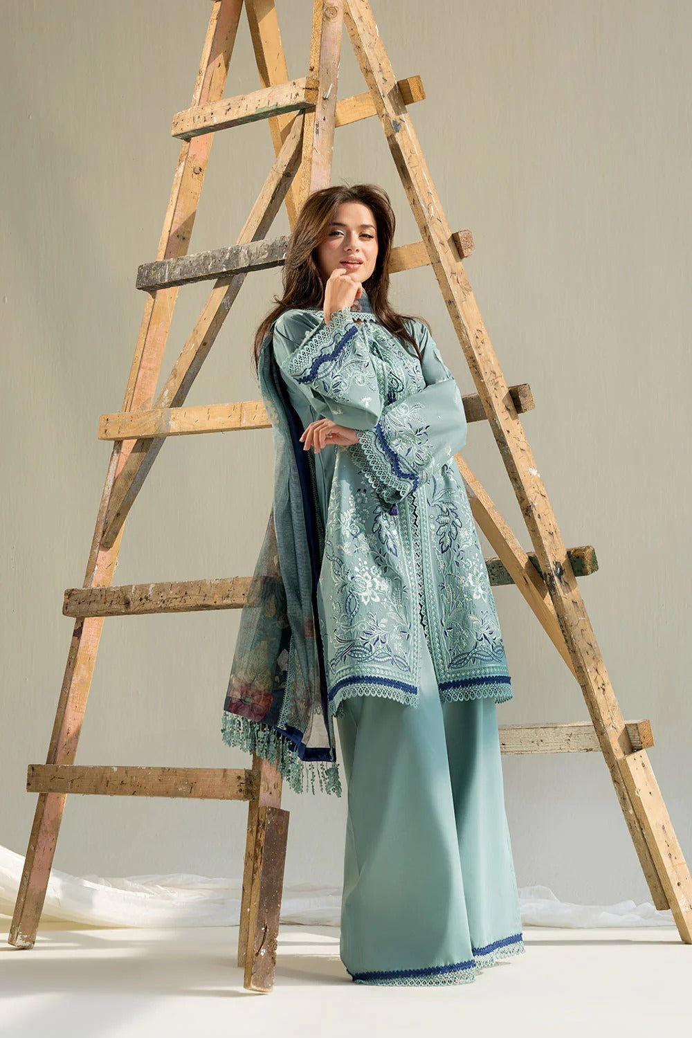 Woman in a light blue embroidered outfit standing next to a wooden ladder on a plain background