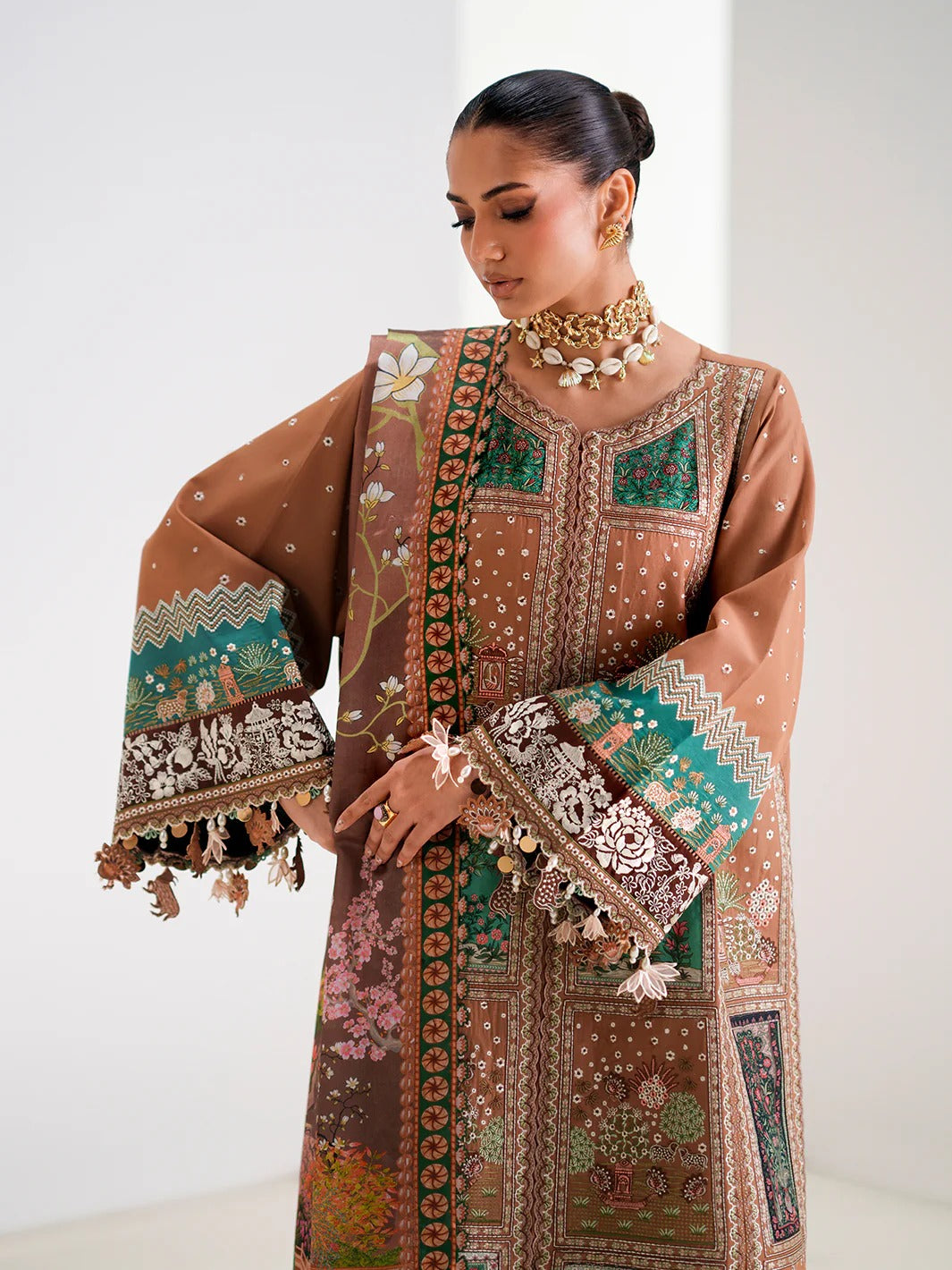 A woman models a brown traditional embroidered kurta set with green and beige patterns, featuring wide bell sleeves with tassels, set against a plain light background.