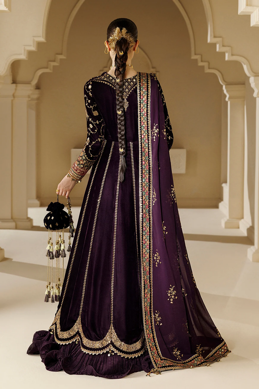 A woman stands with her back to the camera wearing an ornate deep purple traditional outfit with extensive gold embroidery, a matching sheer dupatta, and a black embellished handbag, set against an elegant beige architectural background with arches.