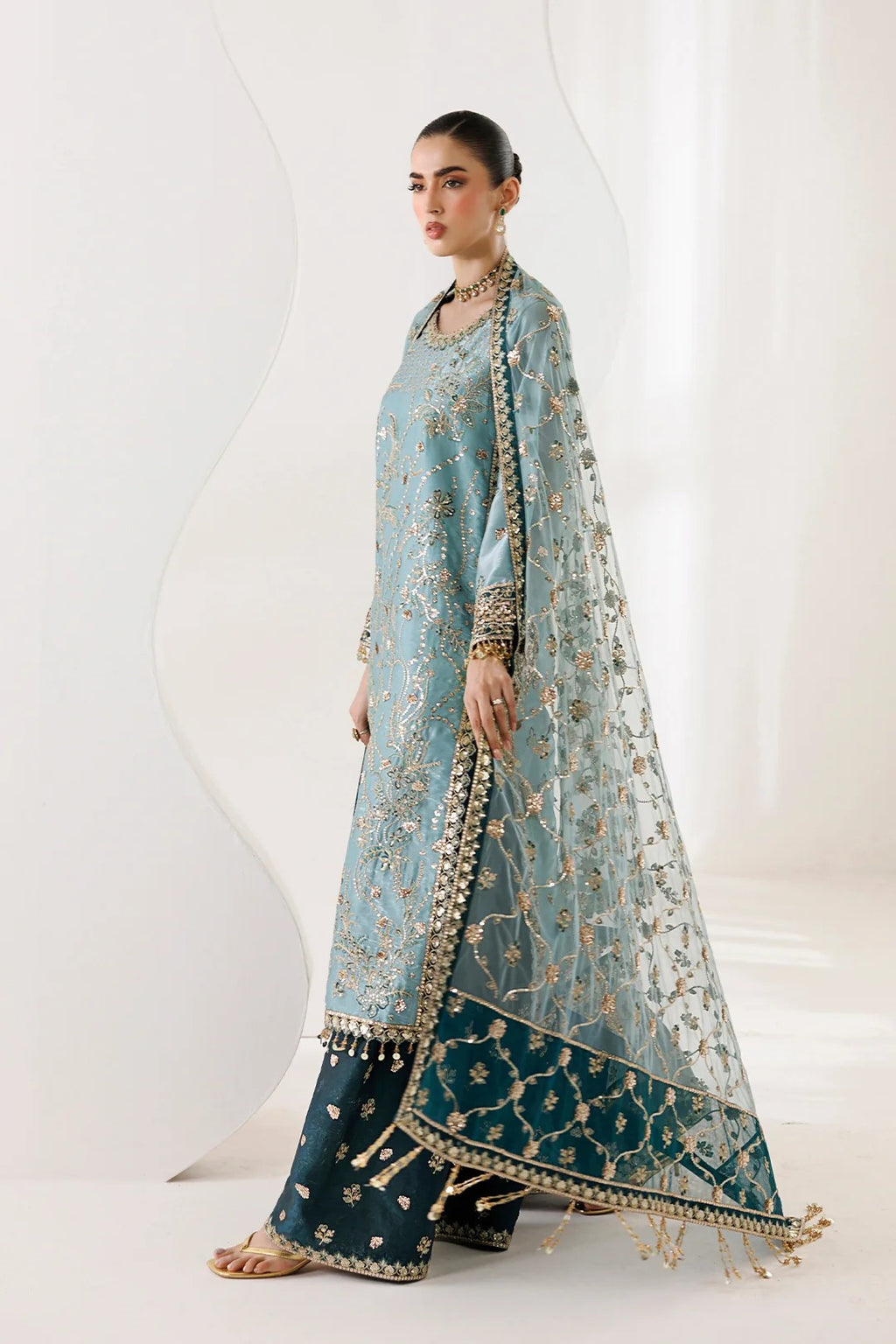 A woman models a traditional South Asian ensemble featuring a light blue embroidered kurta, dark teal wide-leg pants, and a matching sheer dupatta with gold floral embroidery and a decorative border, set against a minimalist white studio background.