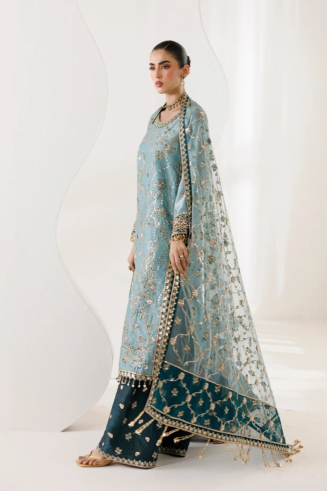 A woman models a traditional South Asian ensemble featuring a light blue embroidered kurta, dark teal wide-leg pants, and a matching sheer dupatta with gold floral embroidery and a decorative border, set against a minimalist white studio background.