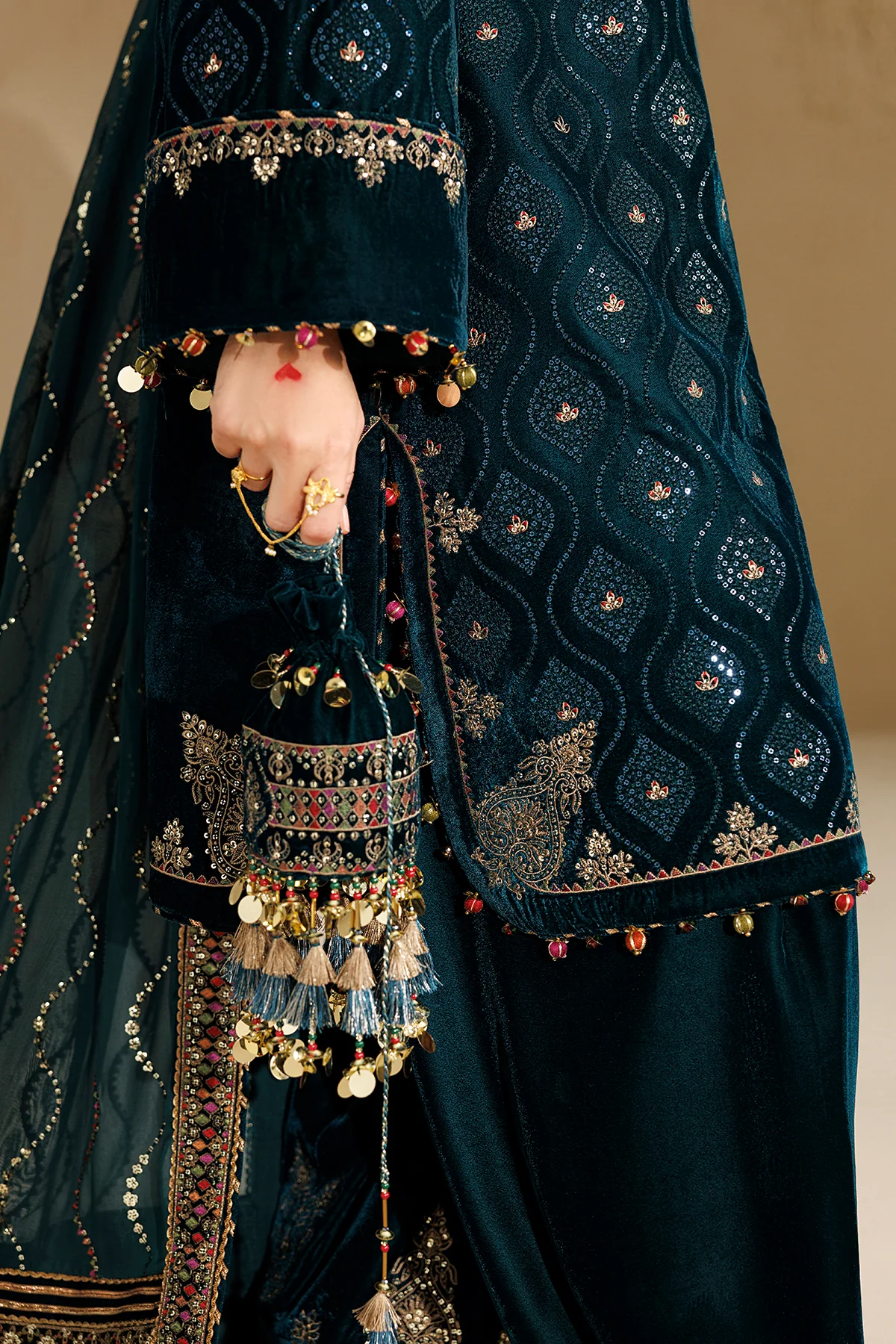 A close-up of a dark teal embroidered traditional outfit with a matching dupatta, featuring intricate metallic embroidery, gold coin tassels, and a hand adorned with rings holding the border.