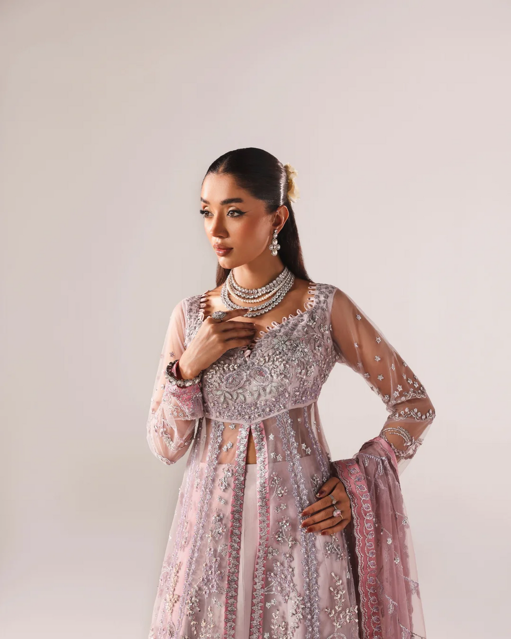 A woman modeling a light pink heavily embellished traditional outfit with silver embroidery, sheer sleeves, and a matching dupatta, photographed against a plain light gray studio background.