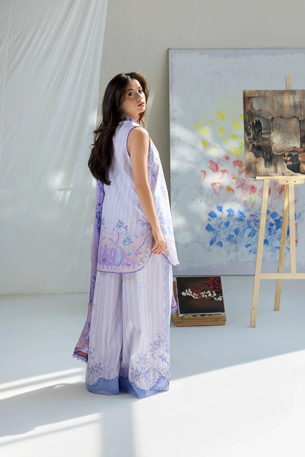 Woman in a light purple outfit standing in a room with a painting on an easel.
